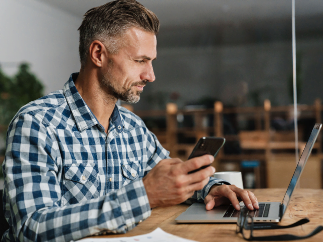 Man sat at desk with phone in hand looking at laptop